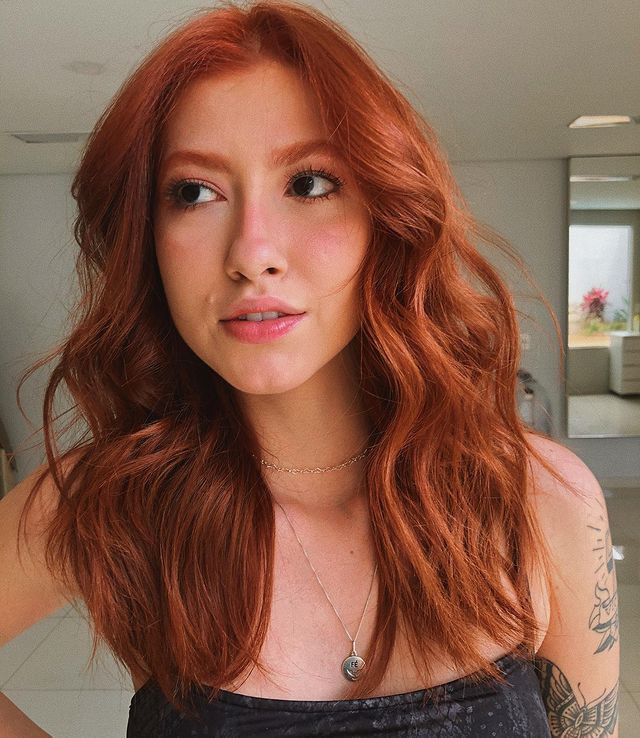 cabelo-ruivo-acobreado Selfie de uma mulher. Ela usa um top cropped preto, cabelo médio ruivo acobreado ondulado com babyliss e maquiagem natural. Ela olha para o lado e não sorri para a foto.