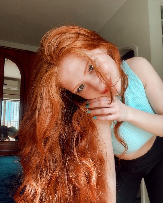 cabelo-ruivo-alaranjado Selfie de uma mulher. Ela usa um top cropped azul, calça legging preta e cabelo ruivo alaranjado longo e solto. Ela olha para a câmera e sorri levemente.