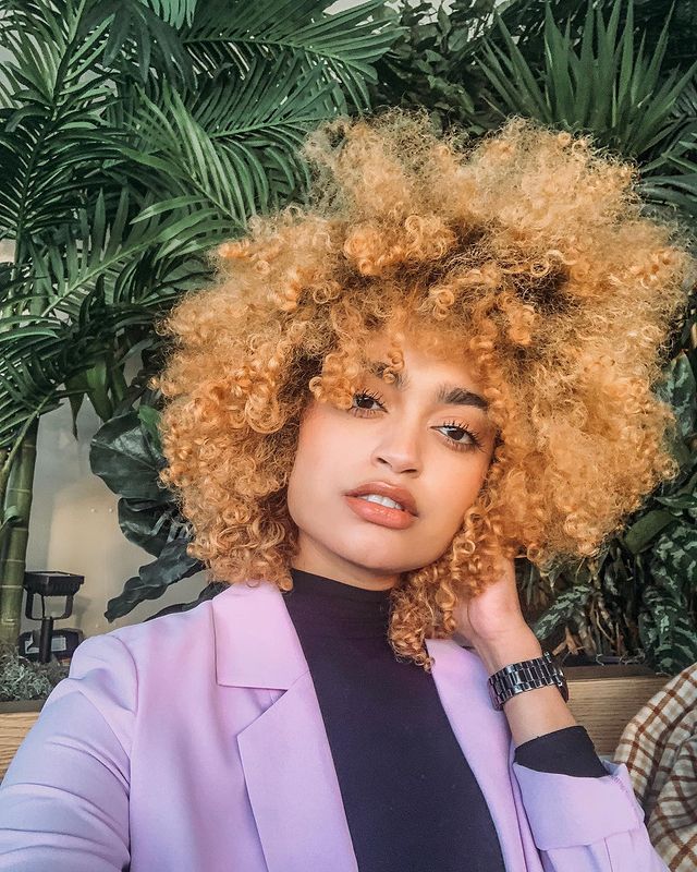 cabelo-ruivo-cacheado Selfie de uma mulher. Ela usa uma camiseta preta de gola alta, casaco lilás como sobreposição, relógio no braço esquerdo e cabelo cacheado ruivo claro solto. Ela olha para a câmera e não sorri.