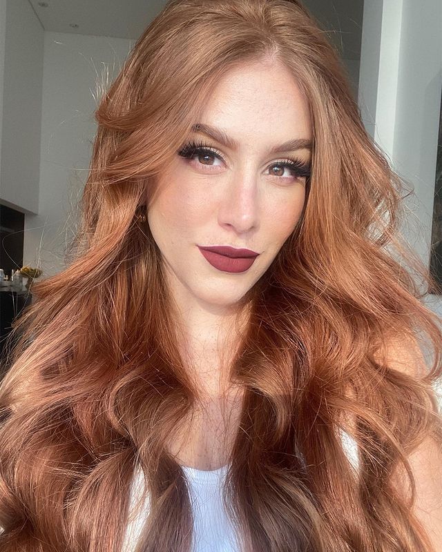 cabelo-ruivo-claro Selfie de uma mulher. Ela usa uma regata branca, cabelo ruivo claro ondulado com babyliss solto e maquiagem com cílios postiços e batom escuro. Ela olha para a câmera e não sorri.