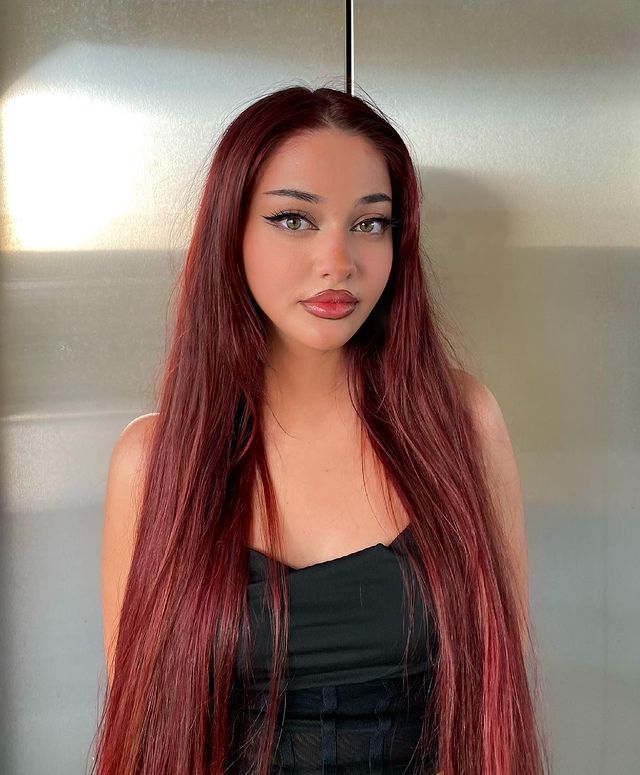 cabelo-vermelho Foto de uma mulher. Ela usa um corset preto, cabelo liso longo e vermelho solto, maquiagem com delineado e gloss nos lábios. Ela olha para a câmera e sorri levemente.