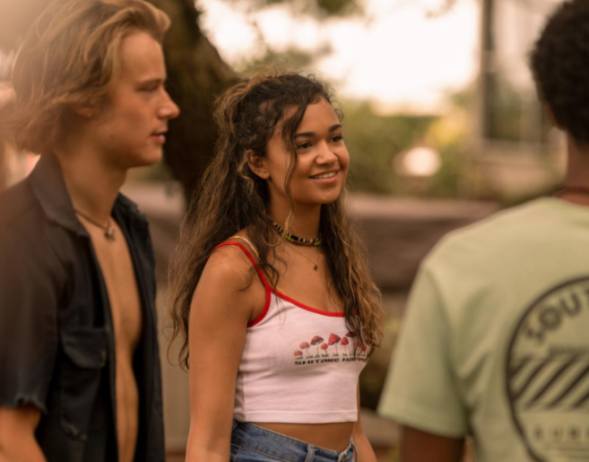 cropped outer banks Três personagens de Outer Banks conversando, um rapaz usa camiseta escura aberta, e menina usa cropped branco e short jeans e o último menino usa camiseta verde, ele está de costas.