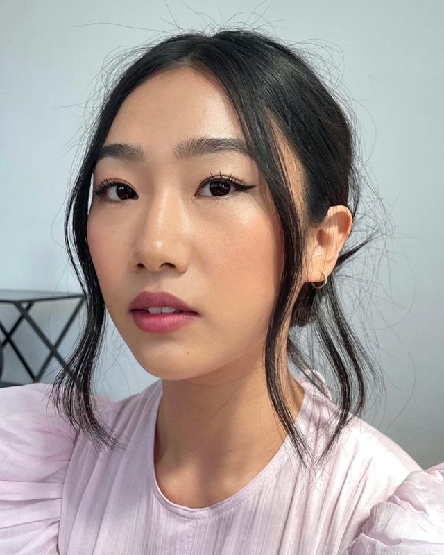 delineado-kitten Selfie de uma mulher. Ela usa uma blusa rosa de manga bufante, cabelo preso em um coque baixo, brinco de argola dourado pequeno e maquiagem natural com mini delineado gatinho. Ela olha para a câmera e não sorri.