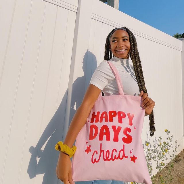 Foto de uma mulher em frente a um muro branco. Ela usa uma blusa de gola alta branca, calça jeans e ecobag rosa com 