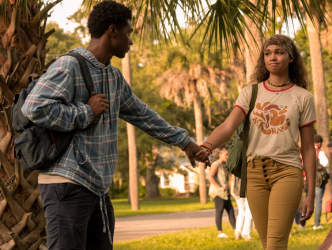 Dois personagens do seriado Outer Banks de mãos dadas em um espaço verde. O rapaz usa blusa azul e calça jeans e a menina camiseta branca com calça bege.