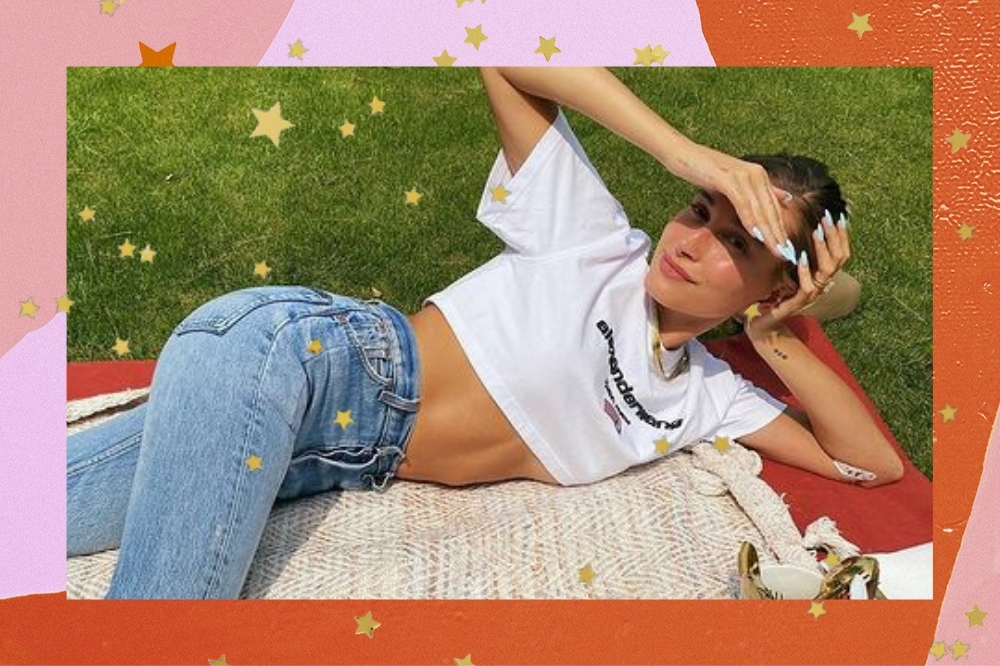 Montagem com o fundo colorido em tons de rosa e laranja com detalhe de estrelinhas douradas com a foto da modelo Hailey Bieber. Na foto ela está deitada em um pano em um gramado, ela usa uma camiseta cropped branca, calça jeans, cabelo preso em um coque e maquiagem natural. Ela está com a mão direita no rosto para proteger os olhos do sol, olha para a câmera e sorri levemente.