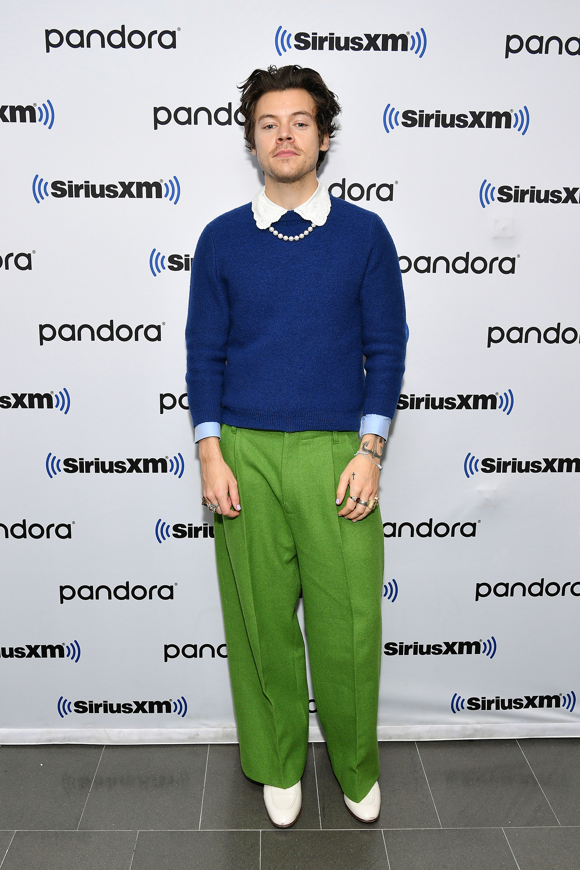 harry-styles Harry Styles usando look com suéter azul e camisa branca com a gola aparecendo, calça pantalona verde e bota branca, além de colar de pérolas. Ele está olhando para a câmera com expressão facial séria.