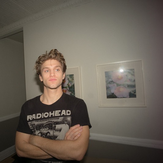 Foto de Keegan Allen com camiseta de banda do Radiohead e os braços cruzados na frente do corpo; ele está com expressão séria e o fundo é uma parede branca com alguns quadros de borda branca pendurados