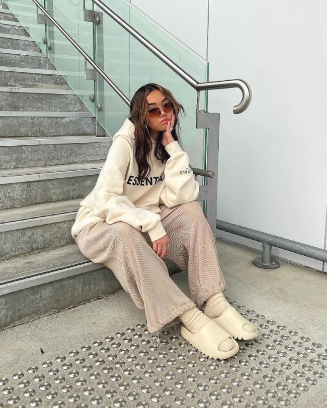 look confy com chinelo nuvem Jovem sentada em escada com expressão séria usando roupas na cor bege.