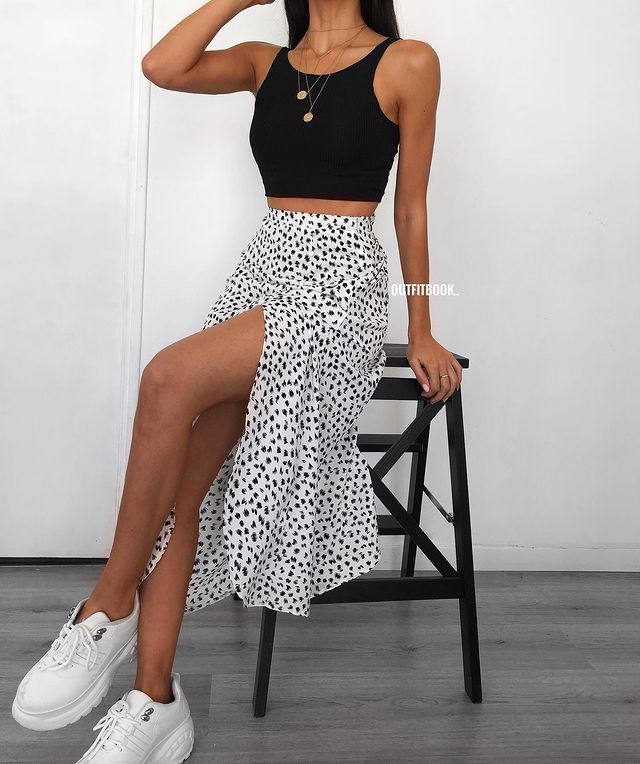 Foto do look de uma mulher sentada em um banquinho preto. Ela usa uma regata preta, saia midi branca com estampa preta, tênis branco e colores dourados.