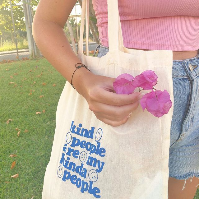 Foto do look de uma menina em um parque. Ela usa um top cropped rosa claro, shorts jeans, ecobag com estampa azul e segura uma flor na mão direita.