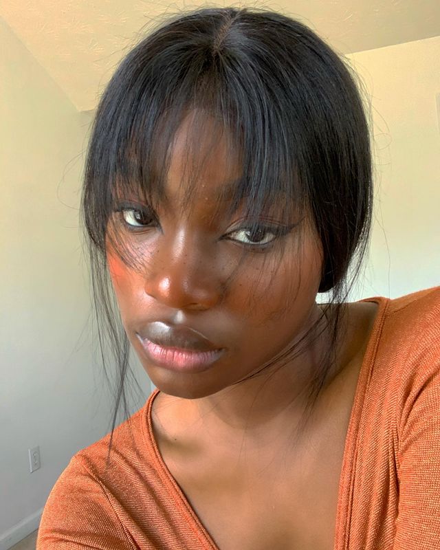 make-lapis-de-olho-preto Selfie de uma mulher. Ela usa uma blusa laranja, cabelo meio preso com franja repicada solta e maquiagem natural com blush marrom, lápis de olho preto na linha d'água e delineado preto. Ela olha para a câmera e não sorri.
