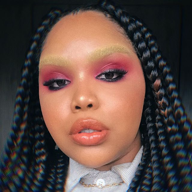 make-rosa-lapis-de-olho-preto Selfie de uma mulher. Ela usa uma camisa, colar dourado, cabelo trançado solto e sua maquiagem é rosa com delineado preto, lápis de olho preto, pele natural, gloss e sobrancelha descolorida. Ela olha para a câmera e não sorri para a foto.