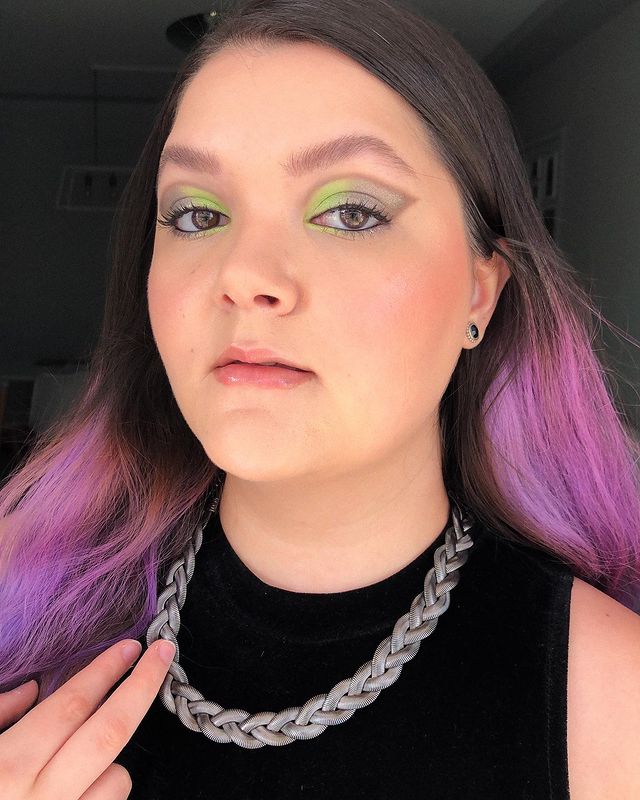 make-verde-lapis-de-olho-preto Selfie de uma mulher. Ela usa blusa preta de gola alta, corrente prata, cabelo solto e maquiagem com pele natural, gloss e olho com sombra verde e cinza metalizada com lápis de olho preto na linha d'água. Ela olha para a câmera e não sorri.