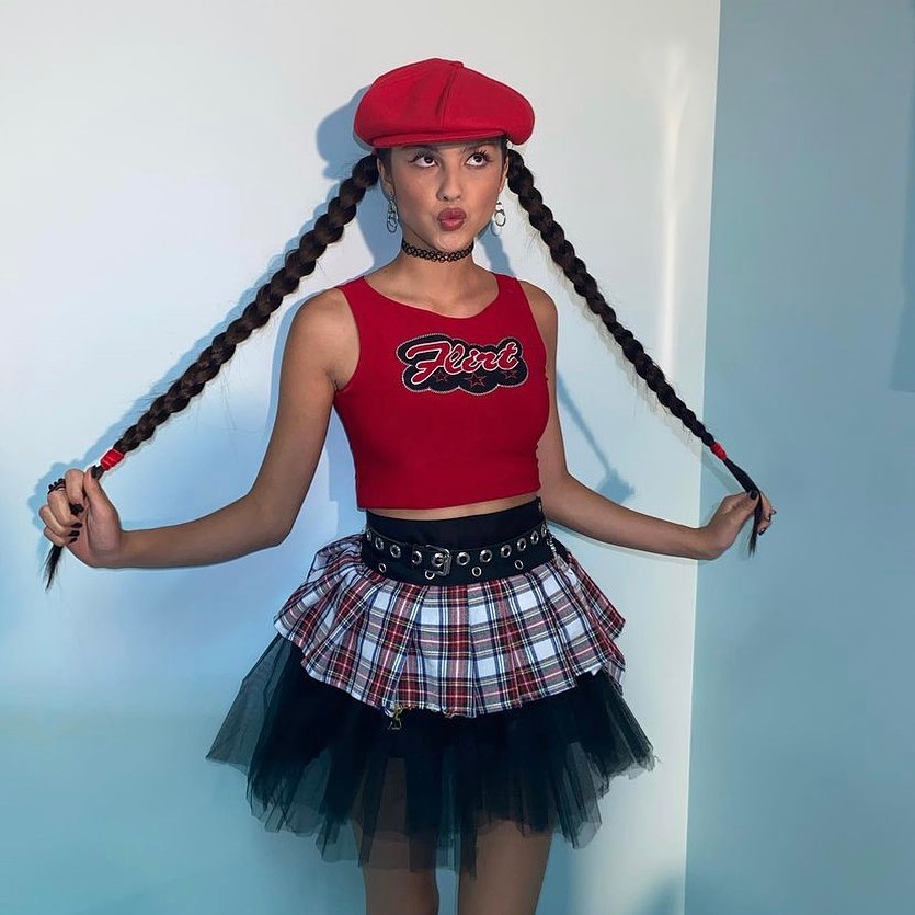 olivia-rodrigo-penteado-claytonhawkins Olivia Rodrigo usando regata cropped vermelha, saia xadrez com tule preto e boina vermelha. Ela está com o cabelo preso em duas tranças laterais, fazendo bico e segurando as tranças com as duas mãos.