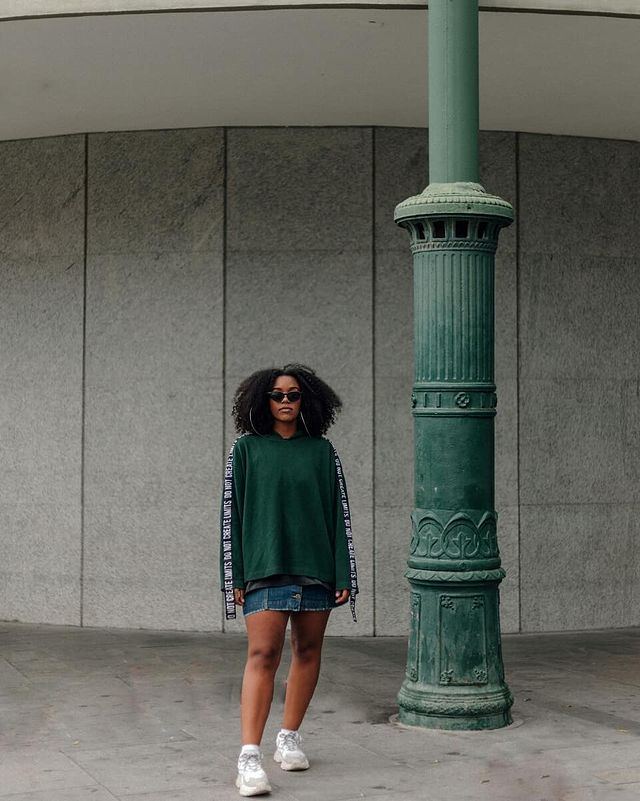 Jovem posando com moletom verde e saia jeans com tênis branco.