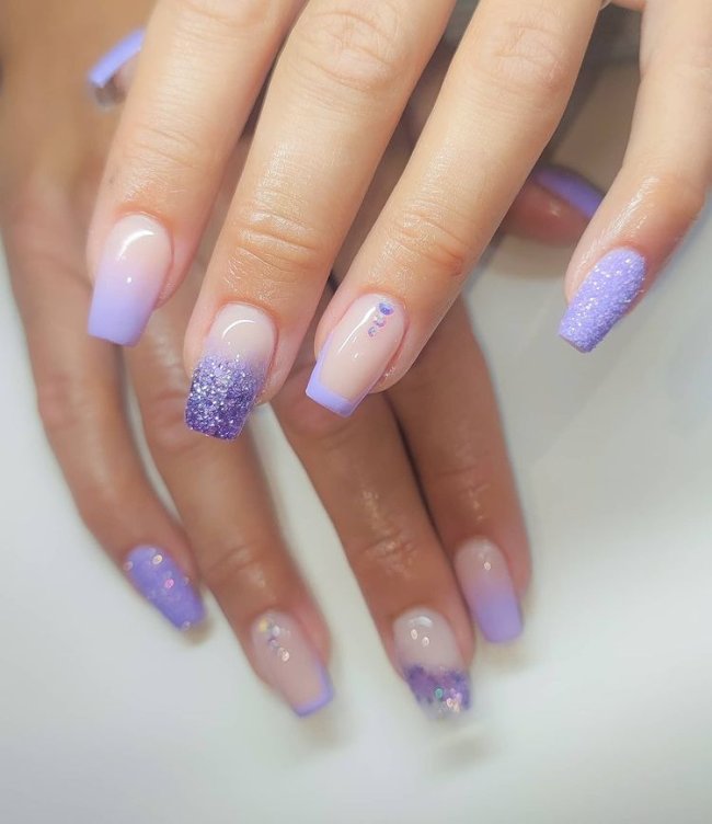 Foto de uma mão com close nas unhas decoradas no estilo baby boomer na cor lilás com detalhes de pedrinhas na unha do dedo anelar.