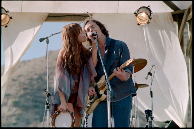 Daisy Jones and The Six – First Look Daisy Jones and The Six; Daisy e Billy cantando no palco e dividindo um microfone enquanto Daisy segura um microfone e Billy toca guitarra