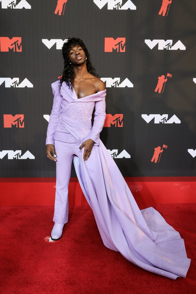 lil-nas-x-mtv-vma-2021 Foto do Lil Nas X no tapete vermelho do MTV Video Music Awards 2021. Ele usa uma blusa lilás com uma calda, calça lilás e sapato na mesma tonalidade. Ele está com o cabelo solto, olha para a câmera e não sorri.