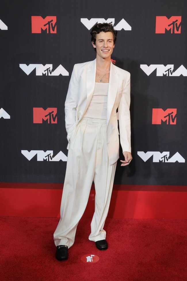 shawn-mendes-mtv-vma-2021 Foto do Shawn Mendes no tapete vermelho do MTV Video Music Awards 2021. Ele usa um terno e calça off white, sapato preto e colar prata. Ele olha para a câmera e sorri.