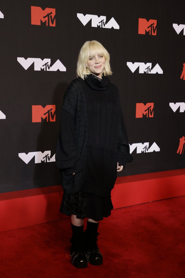 Foto da Billie Eilish no tapete vermelho do MTV Video Music Awards 2021. Ela usa um cardigan oversized preto, saia de renda preta, tênis preto, cabelo solto e maquiagem básica. Ela olha para a câmera e sorri levemente.