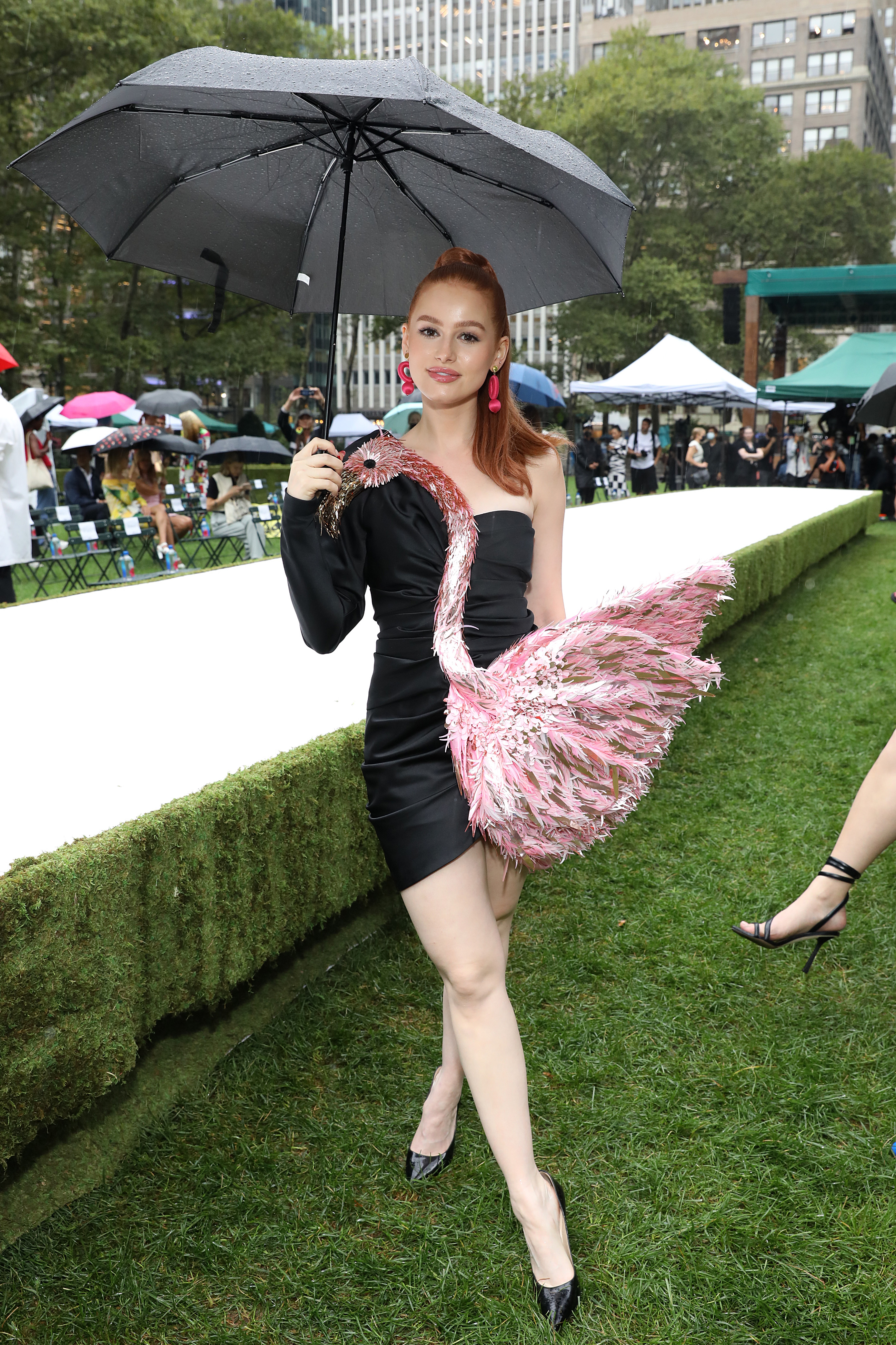 Madelaine Petsch no desfile da Moschino na semana de moda de Nova York. Ela está usando um vestido preto com um flamingo rosa e segurando um guarda-chuva na frente da passarela.