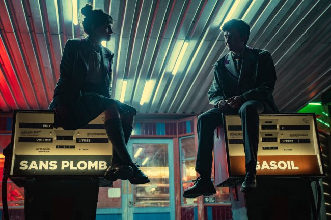 Emma Mackey como Maeve Wiley e Asa Butterfield como Otis Milburn em cena da terceira temporada de Sex Education; eles estão se olhando sorrindo em um posto de gasolina durante a noite; a iluminação vem da loja de conveniência ao fundo e das bombas de gasolina em que estão sentados