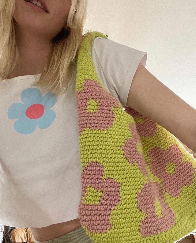 look-com-bolsa-de-croche Foto do look de uma mulher. Ela usa o cabelo solto, blusa branca com estampa de flor e bolsa de crochê verde com florzinhas rosas. A foto não mostra o rosto dela.