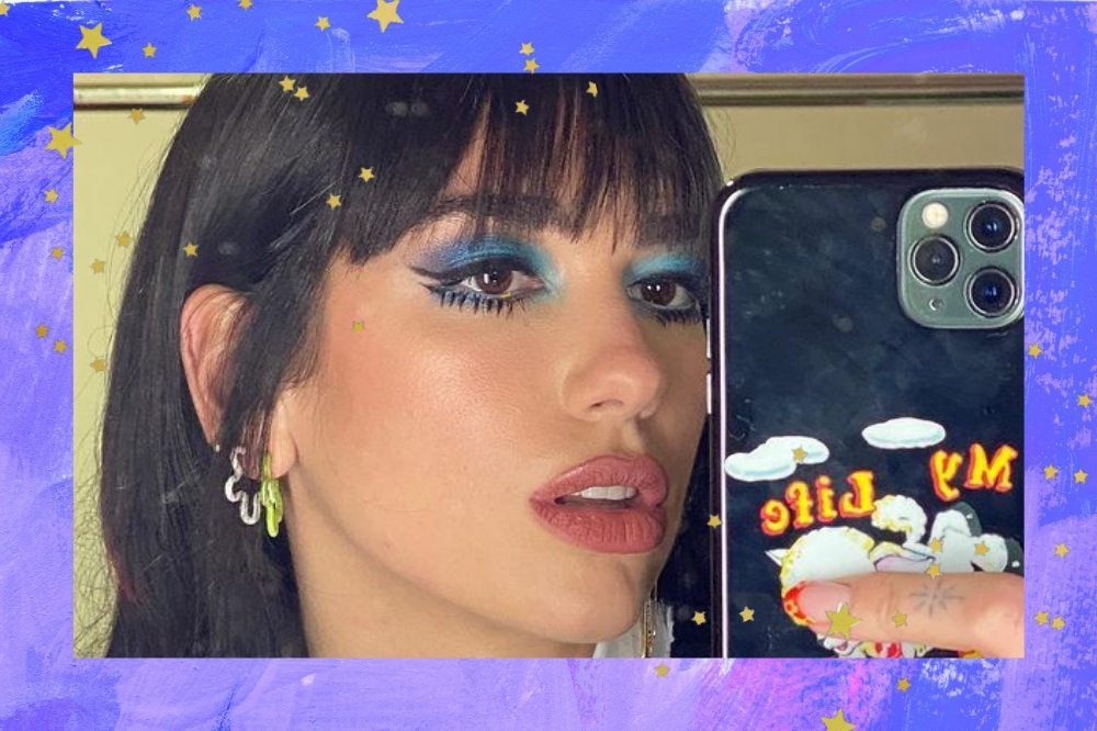 Montagem com o fundo azul e roxo com detalhe de estrelinhas douradas com uma foto da cantora Dua Lipa. A foto é uma selfie da Dua Lipa que está com o cabelo solto com franja, brinco e maquiagem com sombra azul e cílios inferiores destacados.