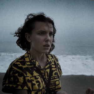 millie-eleven Gif de Millie Bobby Brown como Eleven na série Stranger Things. Ela aparece com blusa estampada amarela e preta em uma praia.