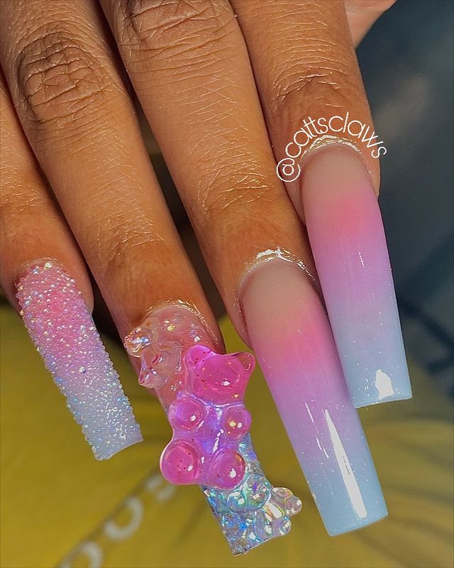 nail-art-com-doces Foto de uma mão com unhas grandes com esmalte degrade azul e rosa claros, o dedo anelar tem decoração de bala de goma de ursinho 3D.