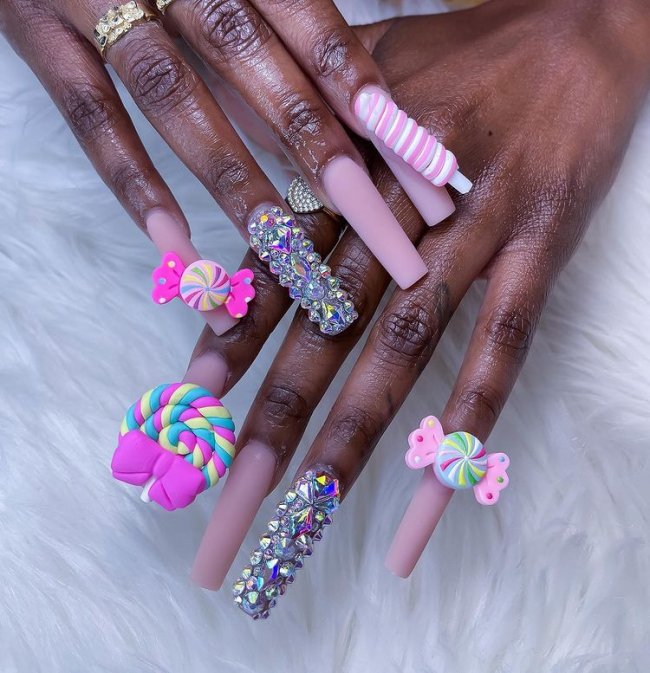 Foto de unhas decoradas. Todos as unhas estão com esmalte rosa claro e detalhes 3D de docinhos coloridos, o dedo anelar está decorados com cristais.