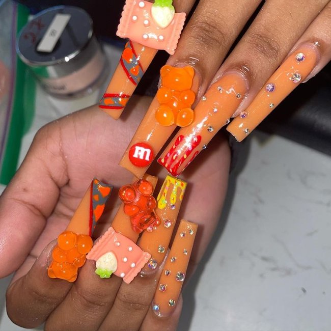 Foto de unhas grandes com esmalte laranja e detalhes de decorações 3D de doces coloridos.