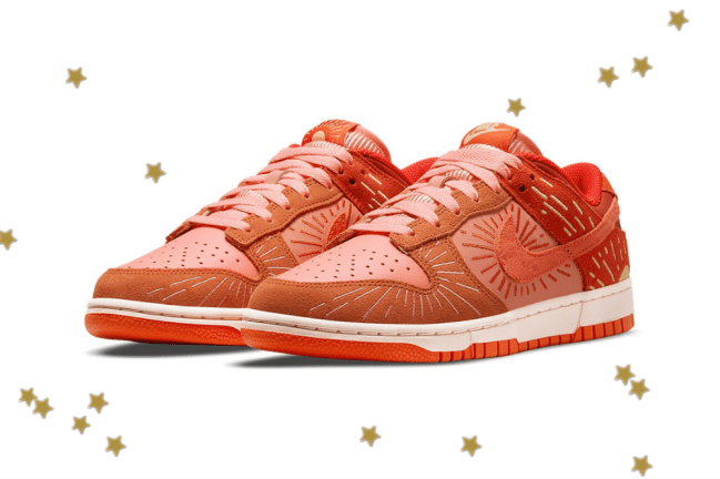 Foto com fundo branco e com estrelinhas douradas. No centro, temos o novo tênis da Nike do modelo Dunk Low com as cores laranja, vermelho e detalhes em amarelo.