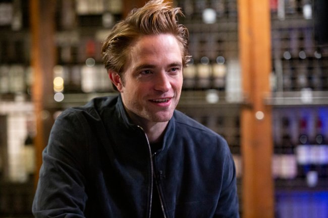robert-pattinson Foto de Robert Pattinson olhando na direção da direita e sorrindo. Ele usa uma jaqueta azul escura e o fundo está desfocado.