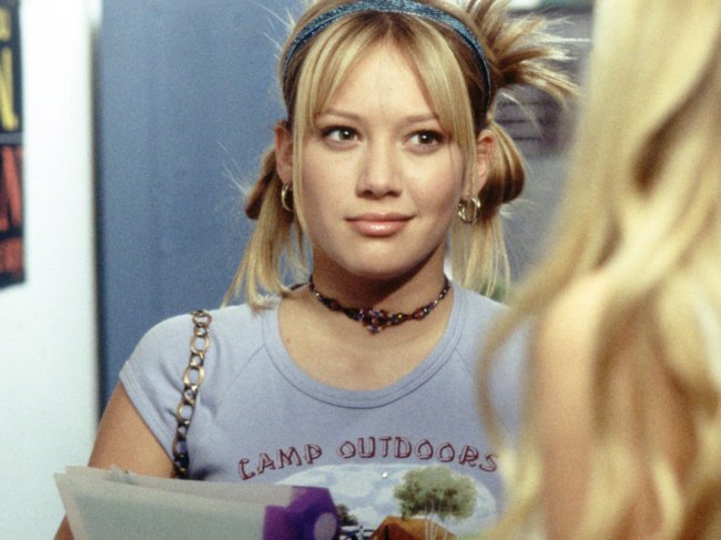 Lizzie Mcguire Hillary Duff como Lizzie McGuire com coques nas laterais do cabelo, tiara e blusa cinza.