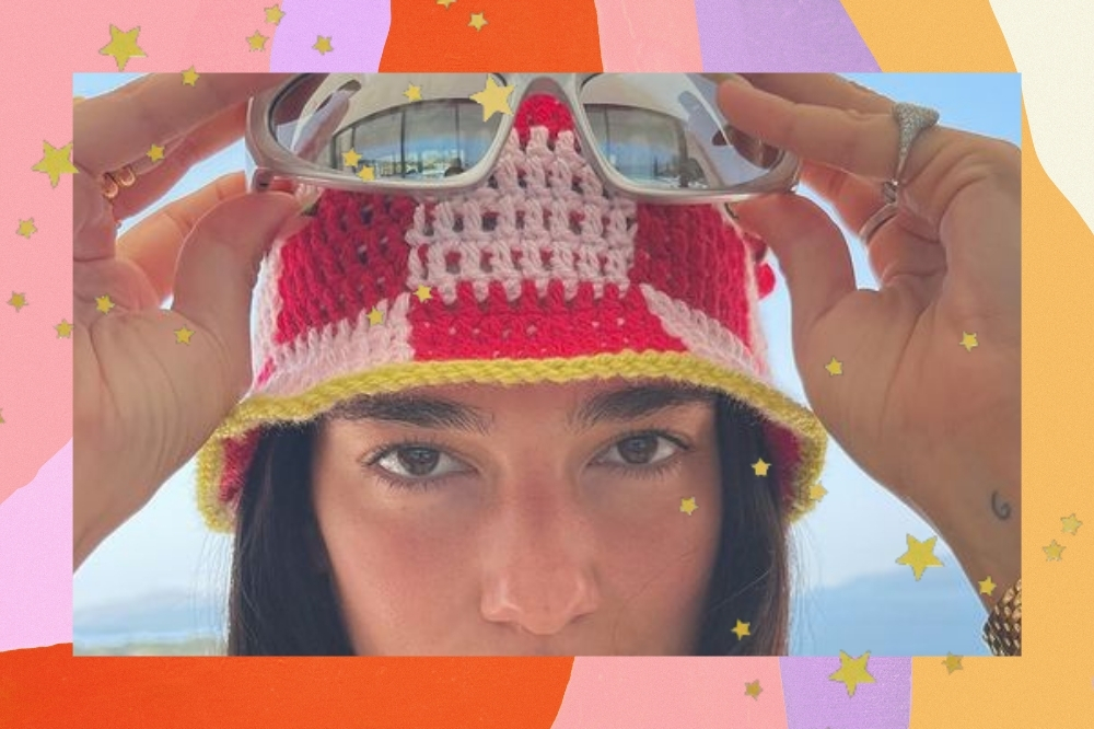 Montagem com o fundo colorido e detalhe de estrelinhas douradas com a foto da cantora Dua Lipa. A foto é um close no rosto dela que mostra ela sem maquiagem, com um chapéu de pescador de crochê colorido e as mãos dela para cima segurando um modelo de óculos metalizado próximo ao chapéu. Ela olha para a foto e não sorri.