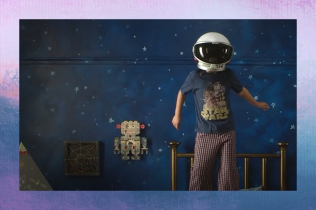 Quarto de Auggie, do filme Extraordinário; ele aparece pulando na cama com camiseta do Star Wars, calça quadriculada em branco e vermelho e capacete de astronauta na frente da parede azul escura com desenhos de estrelas; no canto da imagem é possível ver uma cabana e um item de decoração no formato de um rôbo na cor cinza com detalhes vermelhos pendurado na parede assim como um quadro com linhas brancas e detalhes vermelhos; a margem é uma textura degradê de rosa, branco, azul, lilás e roxo