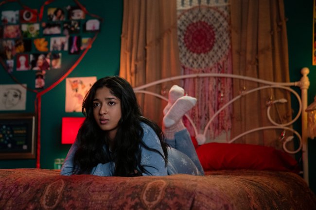eu nunca quarto devi Devi, de Eu Nunca..., série da Netflix deitada na cama de barriga para baixo, olhando para frente com os pés levantados e tem a expressão reflexiva e confusa; ao fundo é possível ver fotos formando um coração na parede verde e um filtro dos sonhos acima da cama e entre cortinas cor de creme; a personagem usa calça jeans e blusa azul