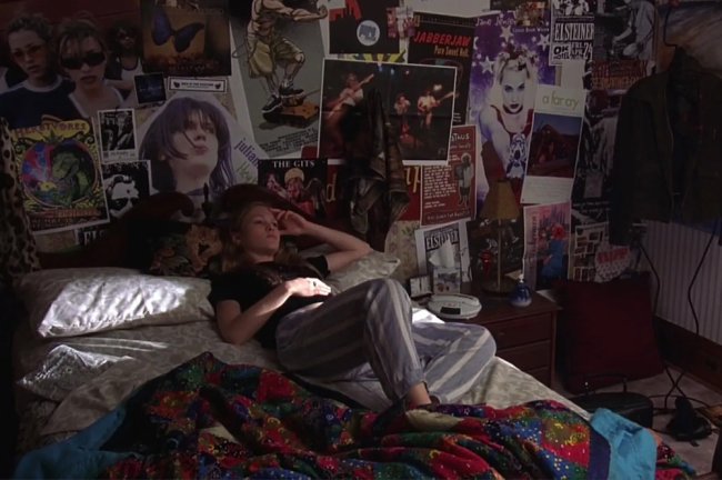 kat 10 coisas que eu odeio em você quarto Quarto de Kat, de 10 Coisas Que Eu Odeio Em Você; a personagem está deitada com a mão apoiada na cabeça vestindo uma calça cinza com listras brancas e uma blusa preta; seu cabelo loiro está solto e ela tem uma expressão séra e reflexiva; a parede do cômodo está preenchida por pôsteres e a iluminação do cômodo vem através da janela