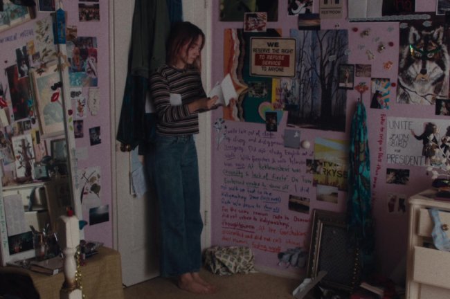 lady bird quarto Quarto de Lady Bird, do filme Lady Bird, produzido pela A24; ela está encostada na porta branca do quarto, que tem roupas penduradas, abrindo um envelope de carta branco com expressão séria; as paredes do cômodo são rosa com pôsteres, frases e desenhos preenchendo o ambiente; um espelho de corpo inteiro está ao lado da porta; a personagem usa calça jeans, blusa listrada e está descalça olhando para os papéis em sua mão