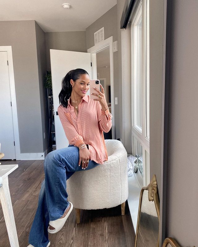 look-calca-e-camisa-colorida Selfie de uma mulher em um espelho sentada no braço de uma poltrona. Ela usa uma camisa rosa de manga longa, calça jeans e chinelo slide. Ela olha para o celular e sorri.