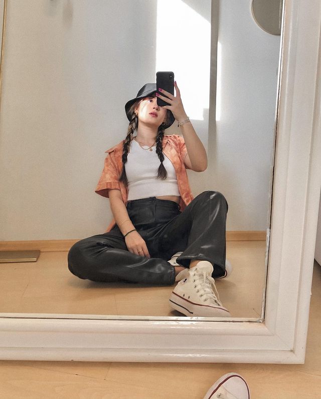 look-camisa-colorida Selfie no espelho de uma mulher sentada no chão. Ela usa uma camiseta branca, camisa laranja tie dye como sobreposição, calça de couro preta, tênis branco de cano alto e bucket hat. Ela olha para o celular e não sorri.
