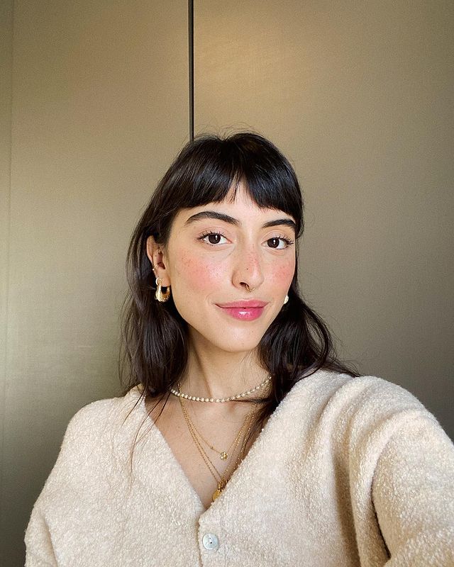 maju-trindade-baby-bangs Selfie da Maju Trindade. Ela usa um cardigan creme, maquiagem bpasica e cabelo liso solto com franja curtinha. Ela olha para a câmera e não sorri.