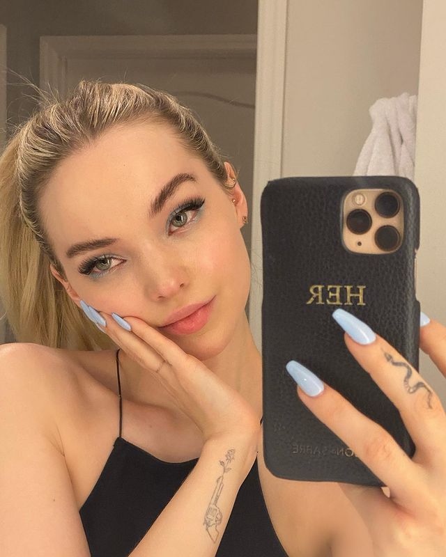 Selfie no espelho da Dove Cameron. Ela usa uma blusa preta, cabelo preso em um rabo de cavalo e maquiagem natural com sombra verde. Ela está com o rosto apoiado na mão direita, olha para o espelho e não sorri.