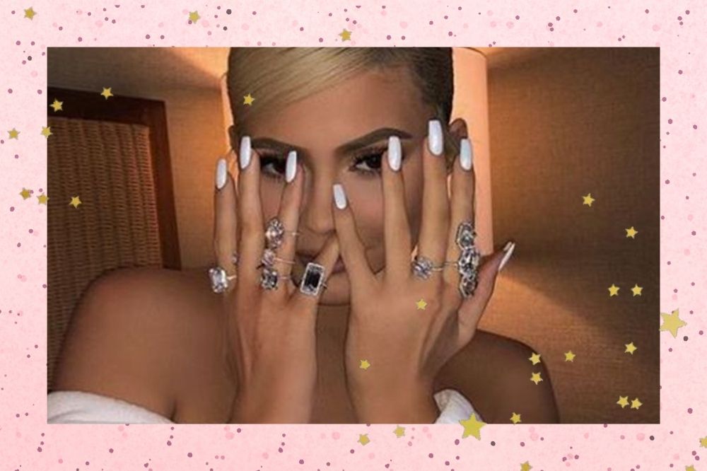 Montagem com o fundo rosa com bolinhas pretas e detalhe de estrelinhas douradas com a foto da Kylie Jenner. Na foto ela está com o cabelo preso e as mãos com a unha pintada de branco na frente do rosto, todos os dedos estão com anéis pratas. Ela olha para a foto e não sorri.