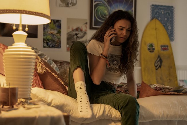 outer banks quarto kiara Madison Bailey como Kiara em cena de Outer Banks; ela está sentada na cama conversando no celular usando uma calça verde com blusa branca e meia branca com detalhes; a parede branca está repleta de quadros e uma prancha amarela está encostada no fundo; um abajur ilumina o cômodo com luz amarela
