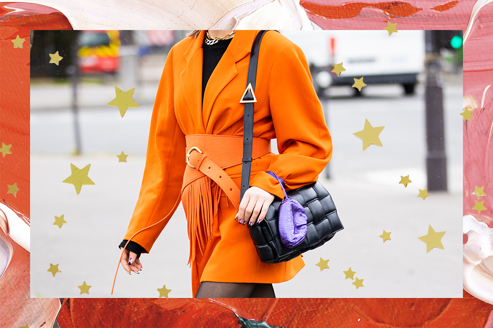 Foto de street style de mulher usando vestido laranja com uma bolsa preta pendurada em um dos ombros. A montagem tem fundo de tintas rosa e vermelha com estrelinhas douradas.