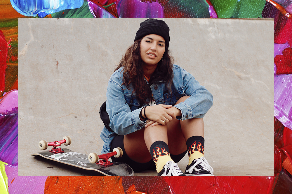 Foto da skatista Thais Gazarra sentada na pista de skate com o skate ao seu lado. A montagem possui fundo de tintas coloridas.