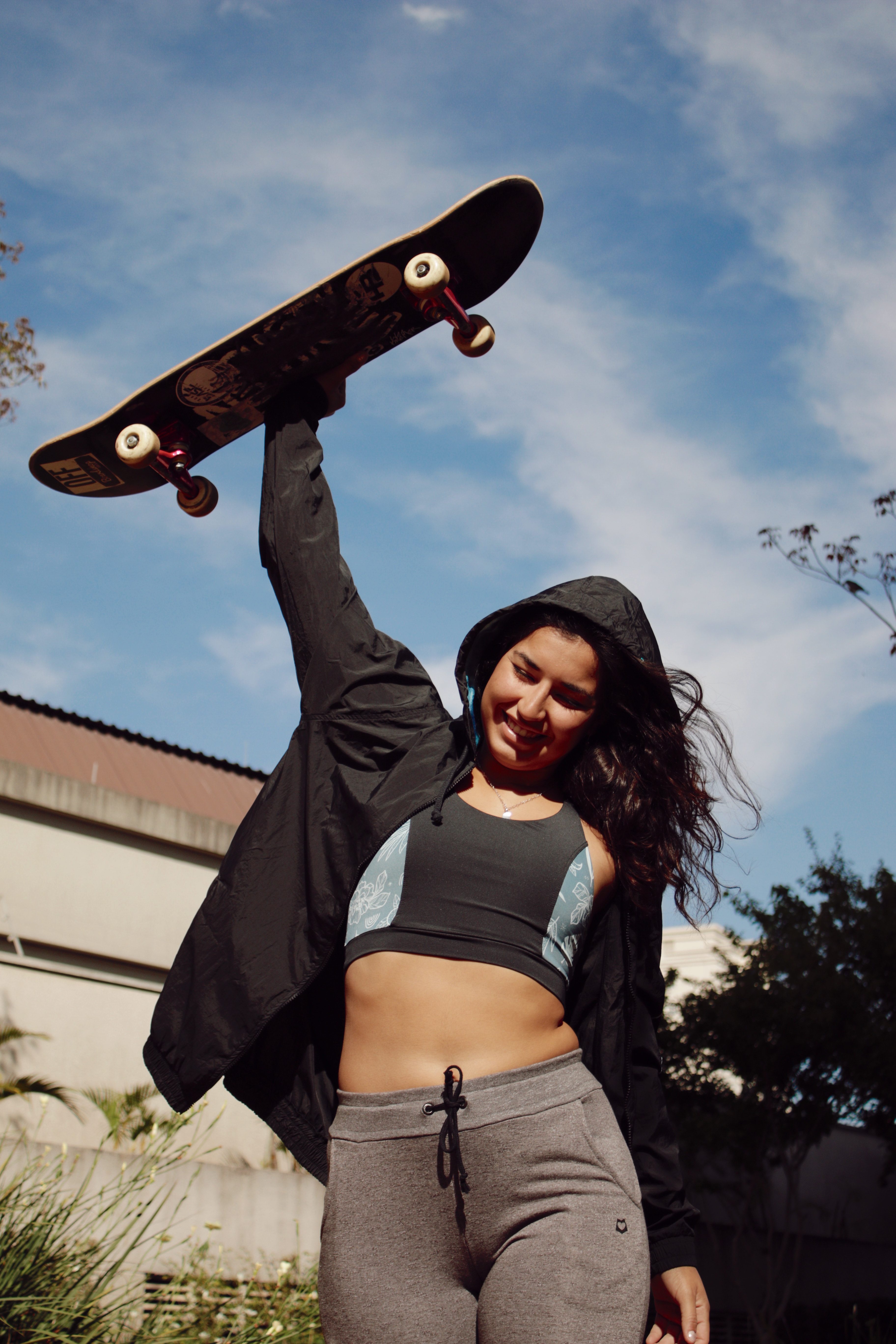 thais-gazarra-skate-48 Thais Gazarra sorrindo, usando um top e uma calça cinza e uma jaqueta preta enquanto segura um skate com uma das mãos e o levanta pra cima